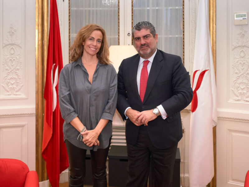 Dos personas posan para una foto formal en un entorno diplomático con banderas y decoración clásica.