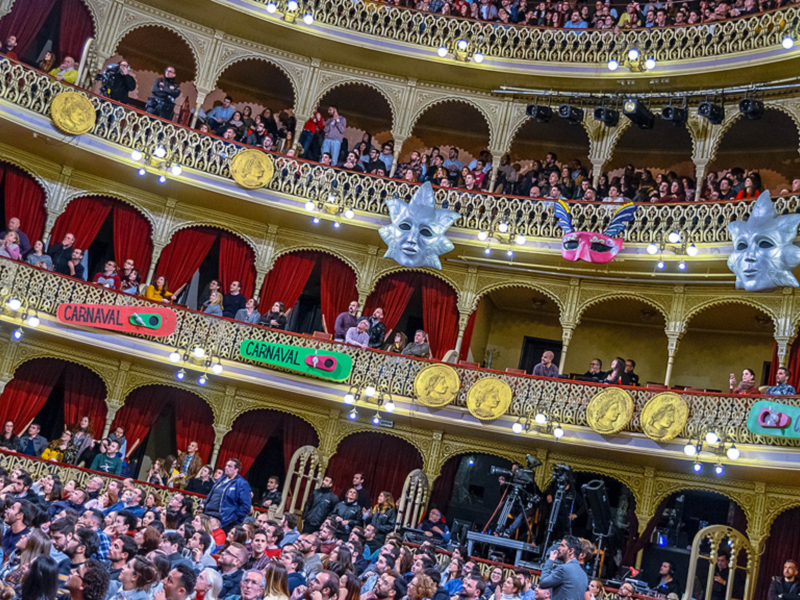 Spectators enjoying a lively carnival event in an ornate theater with colorful masks and festive decorations.