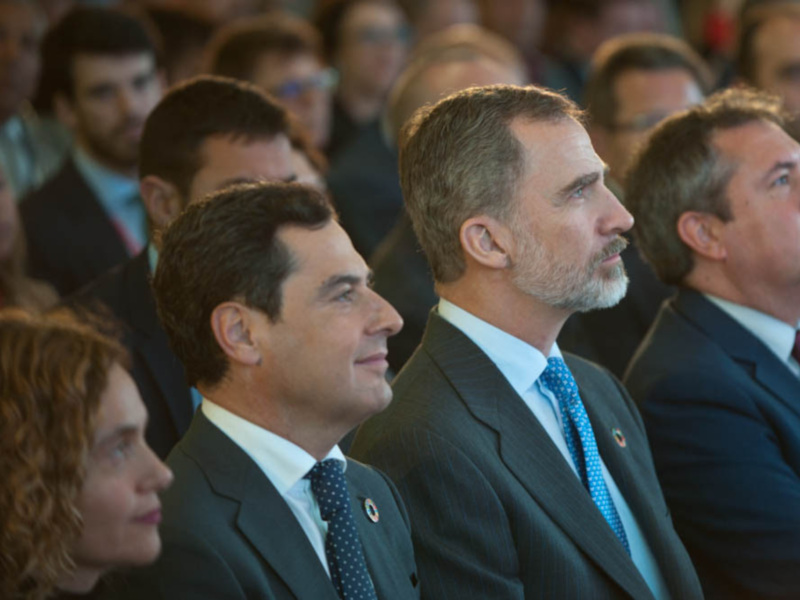 Un grupo de personas en trajes formales, incluyendo a un hombre con una corbata azul con puntos, sonriendo y mirando hacia la derecha.