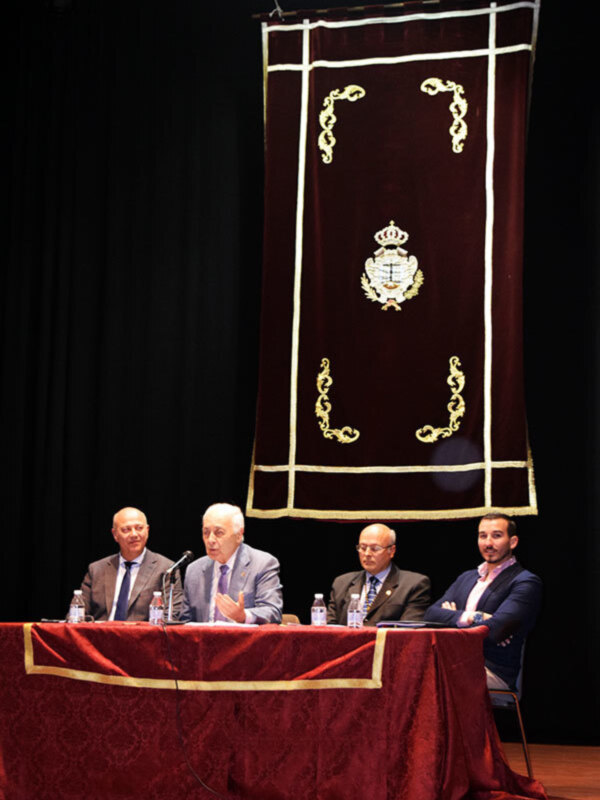 Panel de discusión con cuatro hombres en una mesa bajo un estandarte con escudo realista en un escenario teatral.