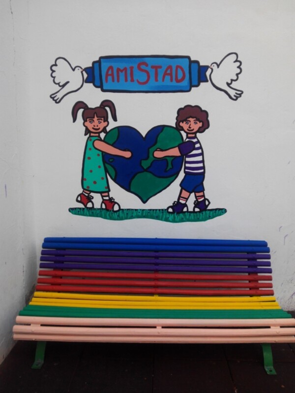 Pintura mural con niños sosteniendo un corazón verde, con la palabra 'AMISTAD' y palomas blancas, junto a un banco de madera multicolor.