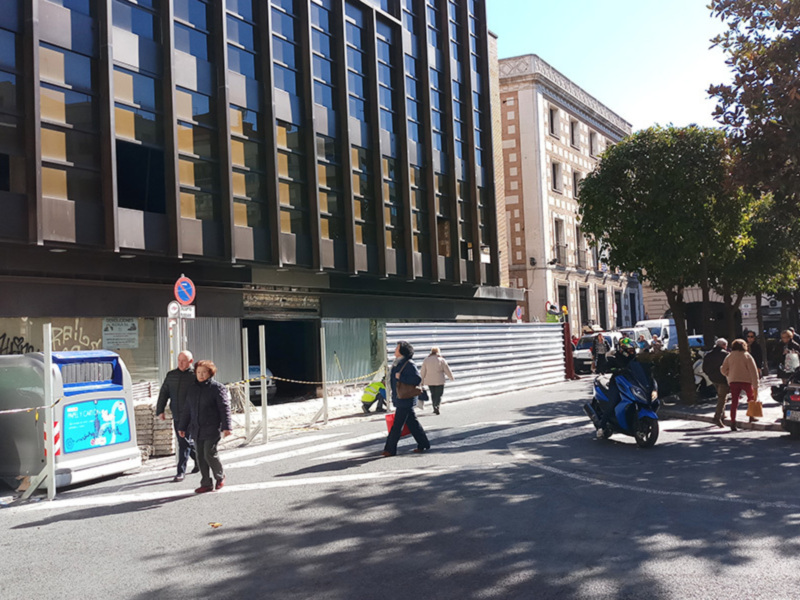 Calle con edificios modernos y antiguos, peatones caminando y motocicletas estacionadas en un día soleado.
