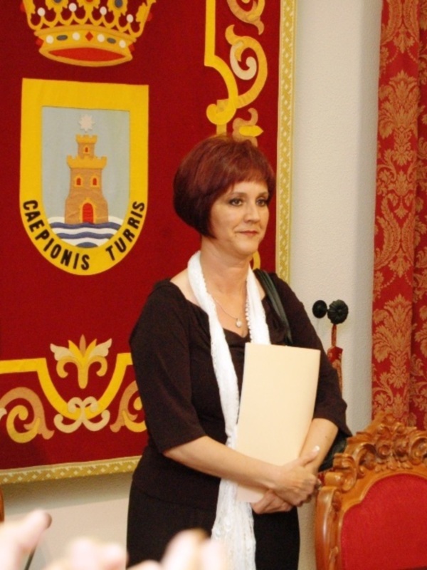 Mujer sonriente en un evento formal, sosteniendo un documento, fondo con escudo y coronas.