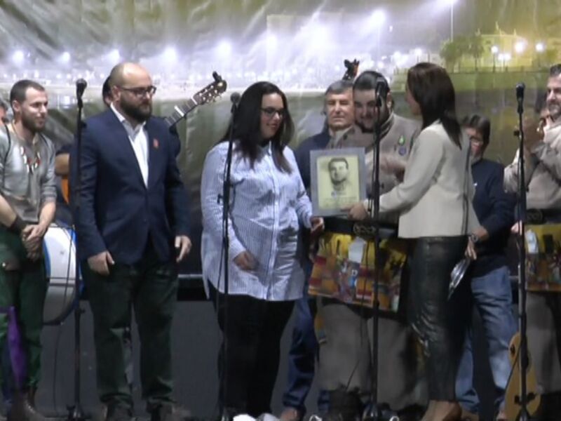 Una ceremonia de reconocimiento en un escenario iluminado, con personas en trajes tradicionales y una mujer sosteniendo un portafoto.