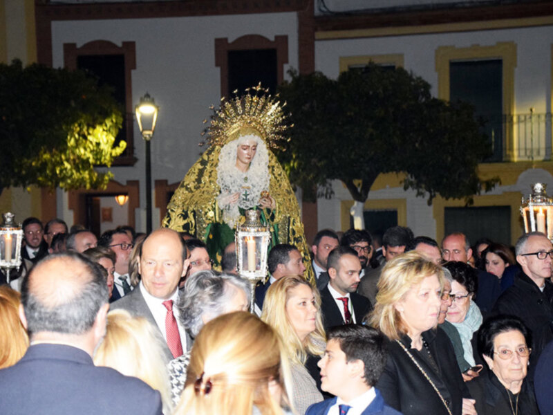 Una procesión religiosa con una imagen venerada en el centro, rodeada por fieles y luces de carnaval. La imagen es la protagonista de la escena, destacándose entre el entorno festivo.