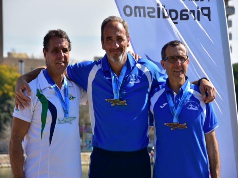 Tres personas posando juntas, dos de ellas con medallas, en un evento deportivo. El fondo muestra una bandera con el texto 'Piragüismo' y un logotipo de una organización deportiva.