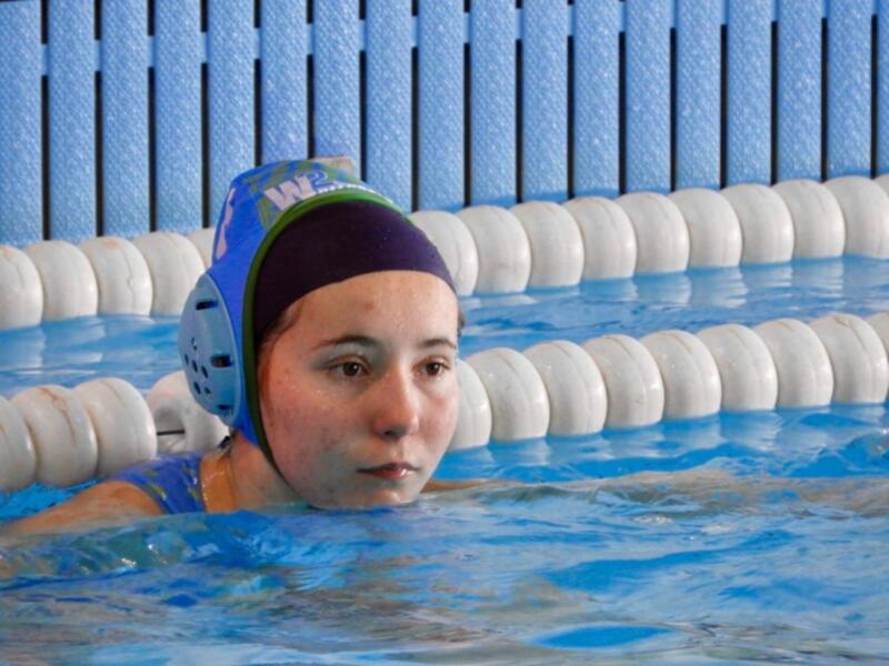 Una joven nadadora en una piscina, con una gorra de natación azul y verde, y una camiseta azul. La piscina tiene azulejos azules y una línea blanca en el fondo.