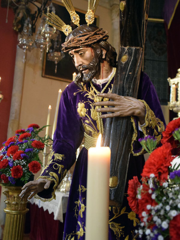 Estatua de Jesús Nazareno en procesión, con túnicas moradas y corona de espinas, rodeada de flores y velas iluminadas.