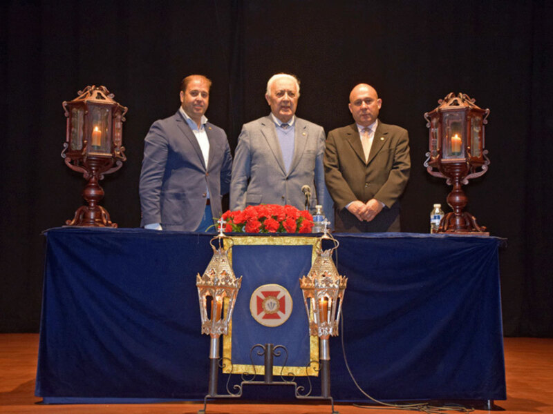 Tres hombres en trajes formales posan detrás de una mesa adornada con candelabros y flores rojas, en un escenario con cortinas negras.