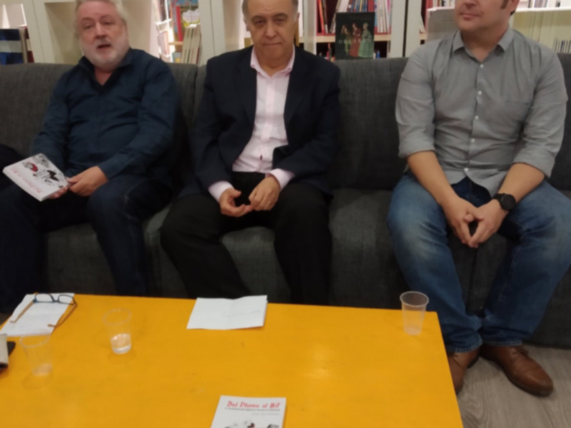 Tres hombres sentados en un sofá en una librería, discutiendo sobre un libro titulado 'Del Diurno al Nocturno'.