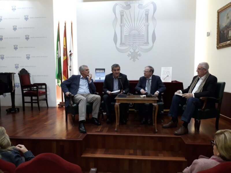 Evento de presentación en el Ateneo de Sevilla con paneles de discusión y banderas de España y Andalucía en el fondo.