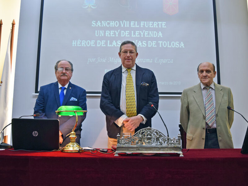 Tres hombres en trajes formales en un evento oficial con banderas y un fondo con texto sobre Sancho VII el Fuerte.