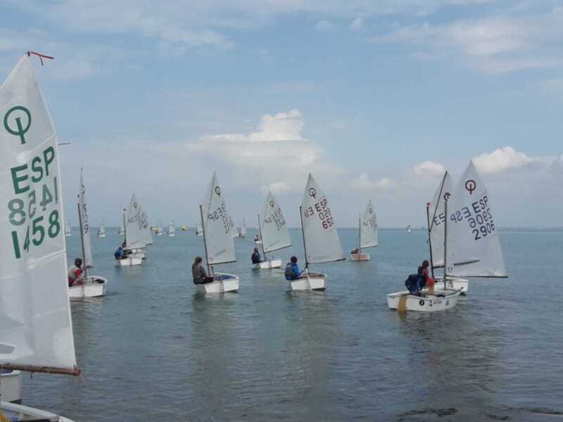 Competencia de vela en barcos Optimist en el mar, con vela blanca y bandera española.