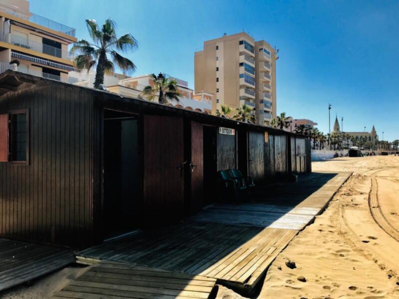 Cabañas de madera en la playa con edificios altos y palmeras al fondo bajo un cielo azul claro.