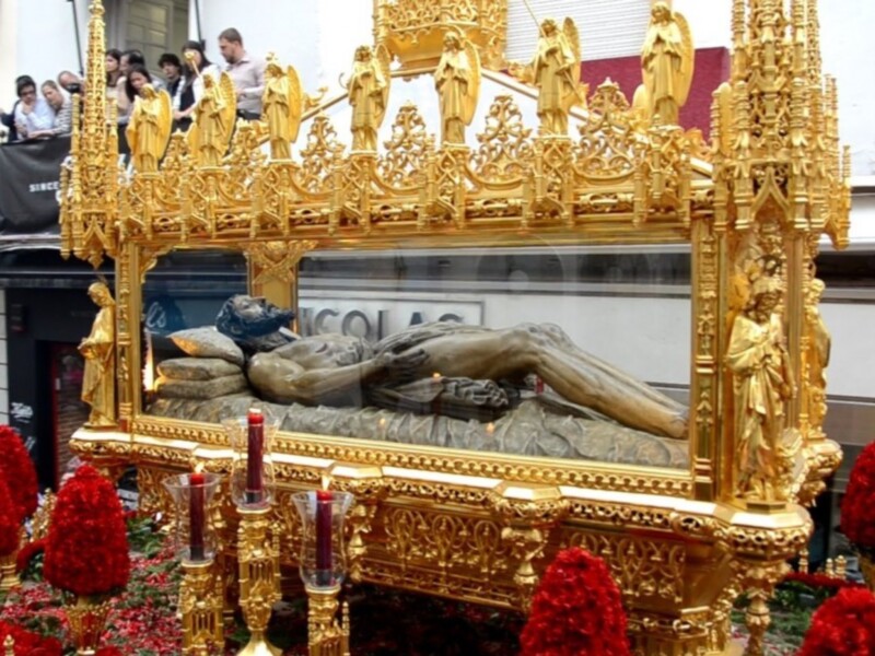 Procesión con estandarte dorado y figura de Cristo en el suelo, rodeado de velas y flores rojas.