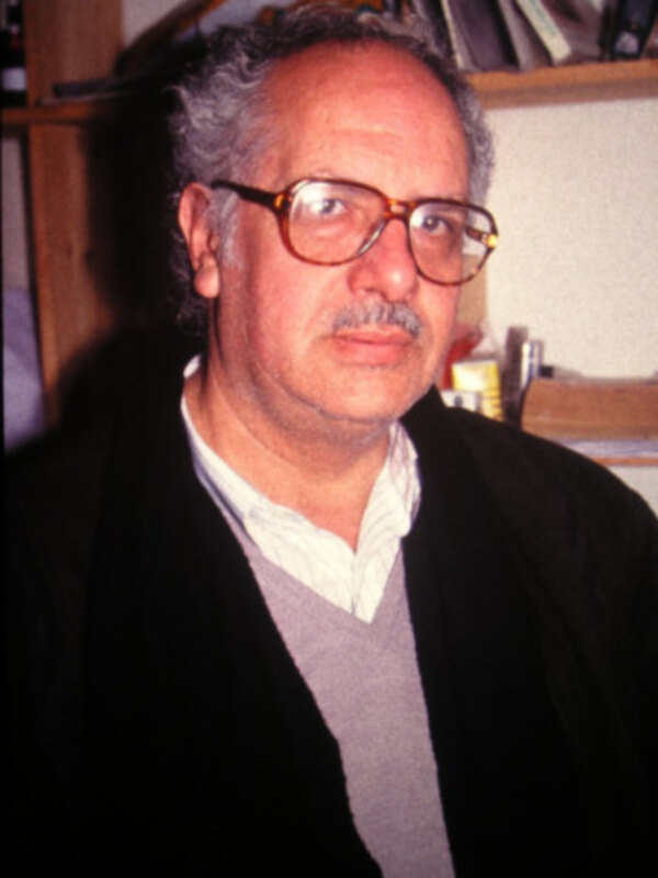 Retrato de hombre con gafas y chaqueta negra, en un entorno interior con estanterías.