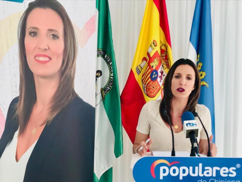 La alcaldesa de Chipiona, María José García, durante una comparecencia ante los medios de comunicación. La imagen muestra a la alcaldesa en un escenario con banderas de Chipiona y España al fondo.