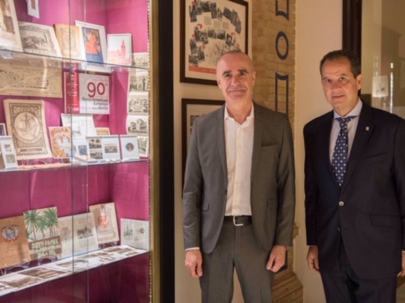 Dos hombres en trajes posan frente a una vitrina con tarjetas postales y libros, en un ambiente cultural.