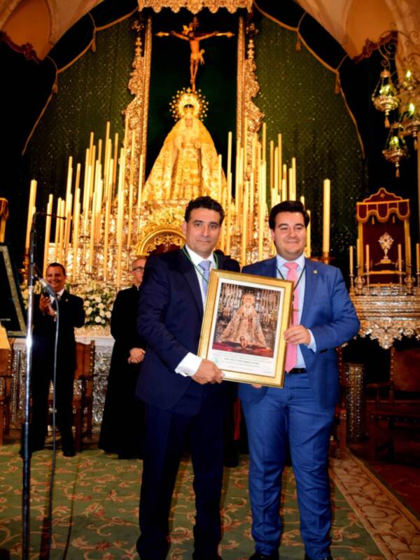 Dos hombres en trajes sostienen un marco con una imagen religiosa en un entorno decorado con velas y una cruz en el fondo.