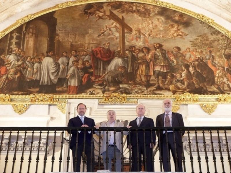 Cuatro hombres en trajes formales posan en un balcón frente a un magnífico lienzo de pintura religiosa, con detalles dorados y figuras históricas en el fondo.