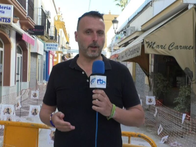 Reportero en una calle comercial hablando al micrófono, con edificios y negocios al fondo.