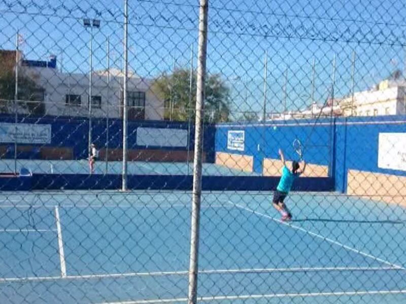 Jugador de tenis en acción en una cancha al aire libre, con una red de alambre y edificios al fondo.