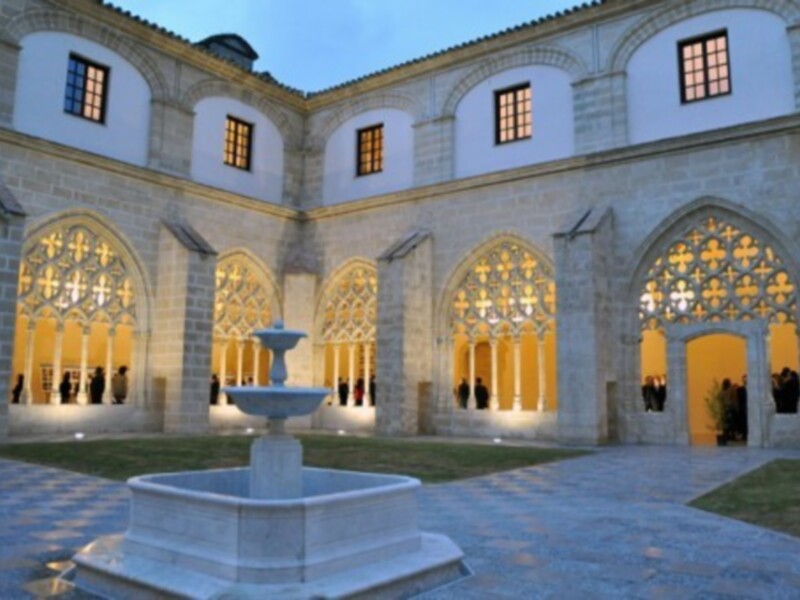 Interior del claustro gótico iluminado por la noche, con arcos y una fuente en el centro.