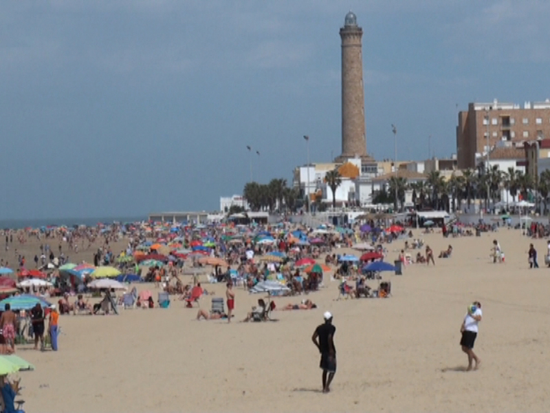 Playa de Muelle Uno, Cádiz, con turistas disfrutando de la playa y el faro en el fondo.