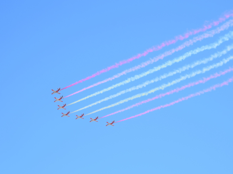 Desfile aéreo con aviones en formación y colores vibrantes en el cielo azul.