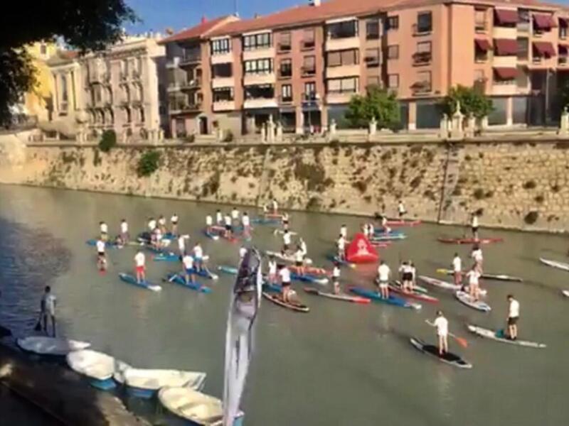 Grup de persones practicant paddle surf a una riera urbana amb edificis al fons.