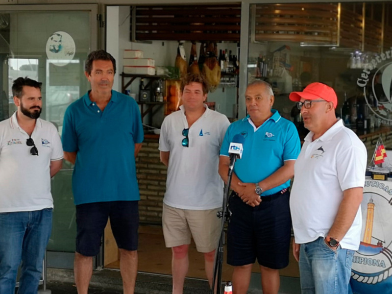 Cinco hombres en uniformes de club náutico posan frente a un micrófono en una presentación o evento.