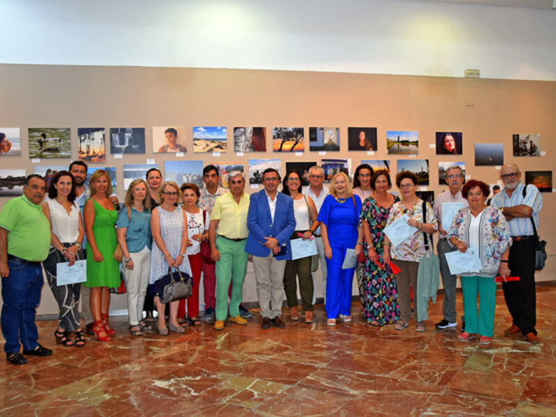 Una reunión de personas en una exposición de fotografías, todos sonrientes y sostienen sus diplomas.
