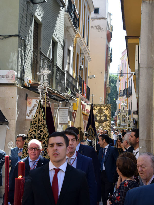 Procesión religiosa con portadores de candelabros y estandartes en una calle urbana.