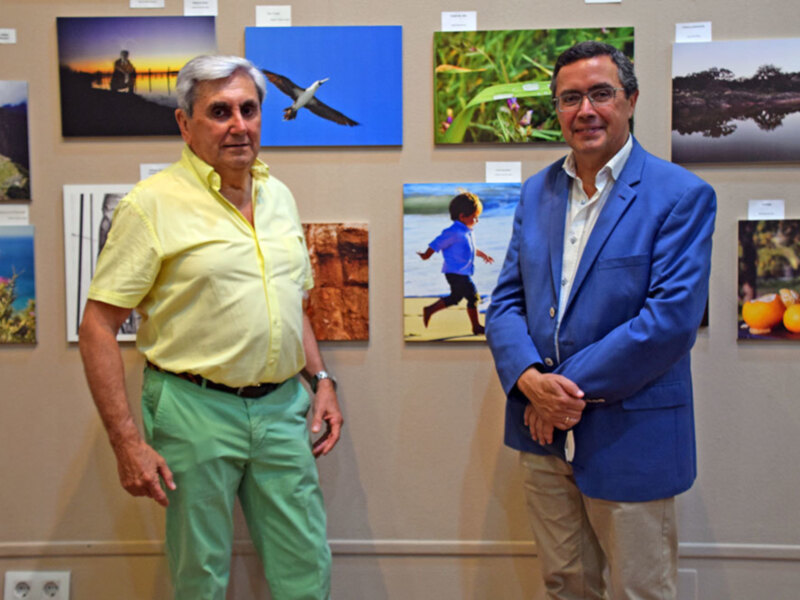 Dos hombres posan frente a una exposición de fotos de paisajes y naturaleza.
