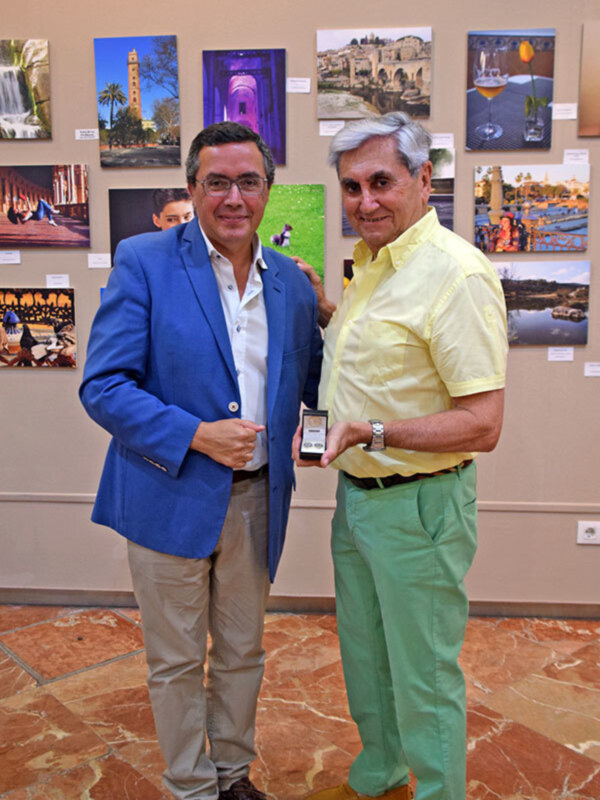 Dos hombres posan juntos en una galería de arte, uno sostiene un teléfono móvil y sonríen hacia la cámara.