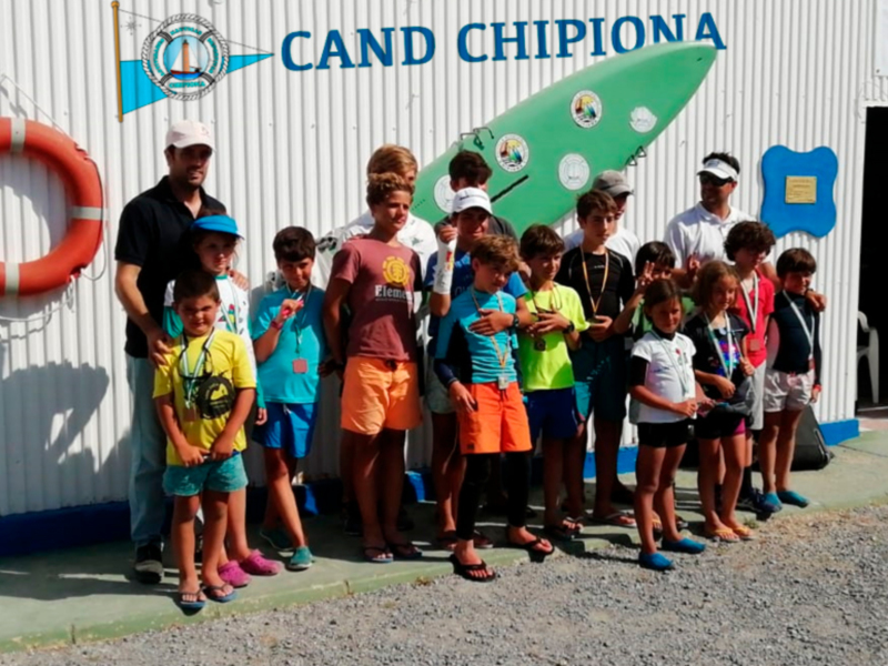 Niños y jóvenes posando con un surf en el Club de Surf Chipiona, con una bandera y una placa en el fondo.