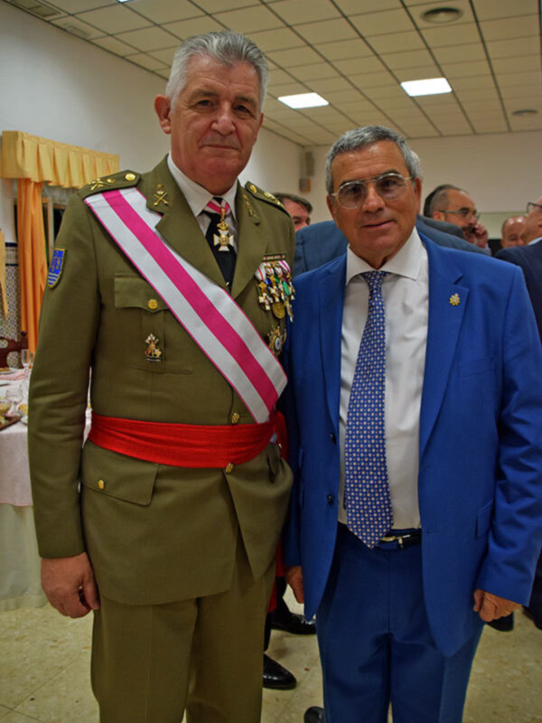 Dos hombres, uno en uniforme militar con sash rojo y otro en traje azul, posan juntos en una recepción formal.