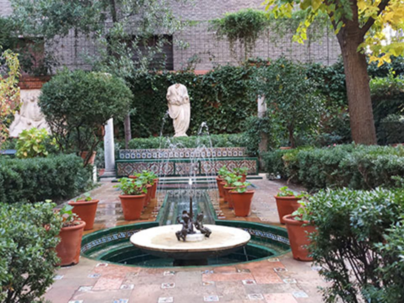 Jardín con fuente central, estatuas y macetas de flores en un patio interior con muros de piedra y vegetación.