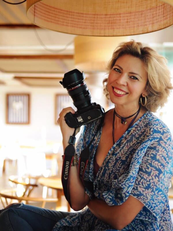 Retrato de una mujer sonriente sosteniendo una cámara Canon, en un ambiente acogedor con lámparas de techo y muebles de madera.