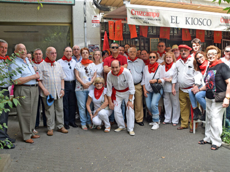 Una multitud de personas se reúne en un evento, con algunos portando bufandas rojas y otros con uniformes blancos y rojos. La imagen muestra una celebración o una reunión social en un entorno urbano, con banderas y carteles en el fondo.