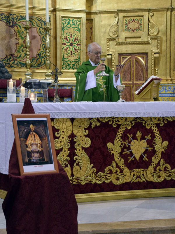 Un sacerdote bendice con un vaso de cristal en una iglesia con una mesa de altar decorada con velas y un cuadro de San Pedro.