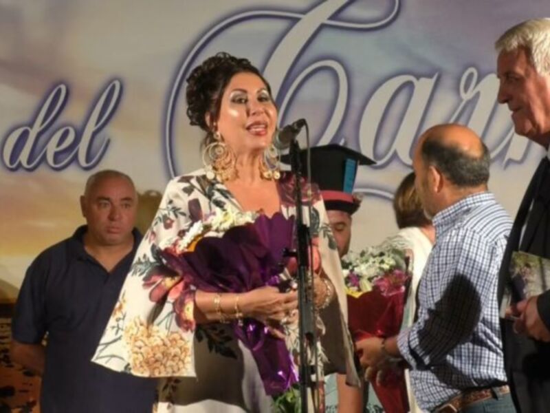Mujer hablando en un evento público con un micrófono, rodeada de personas y un fondo con el texto 'del Caño'.