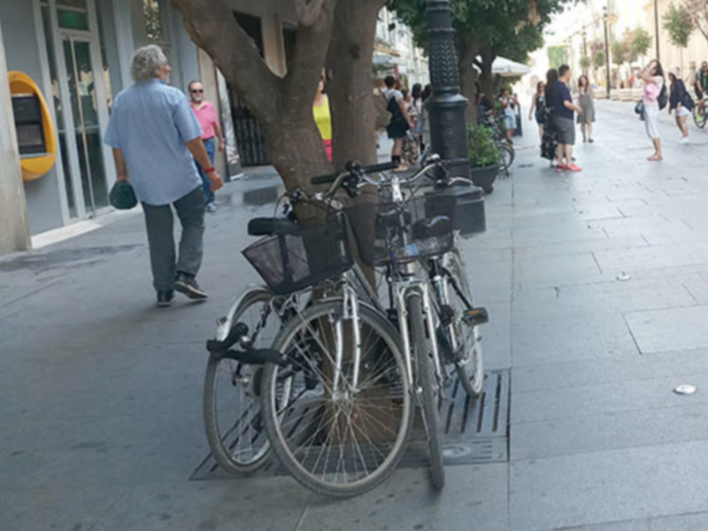 Bicicletas aparcadas en una estación de bicicletas en un entorno urbano con personas caminando en el fondo.