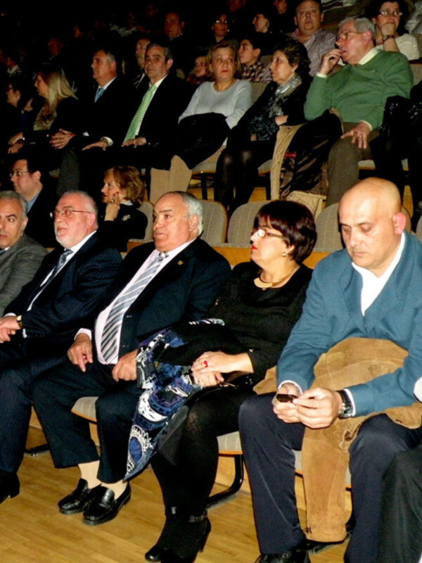 Asistentes a una conferencia o evento, algunos con trajes y otros con ropa casual, sentados en sillas alineadas en una sala de conferencias.