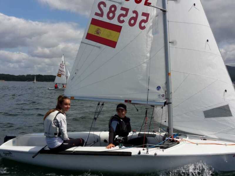 Dos jóvenes en un velero blanco con bandera española, navegando en el mar con otros barcos al fondo.