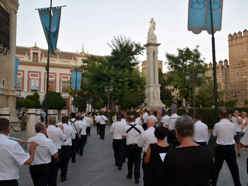 Banda de música desfilando por una plaza histórica con edificios y monumentos al fondo.