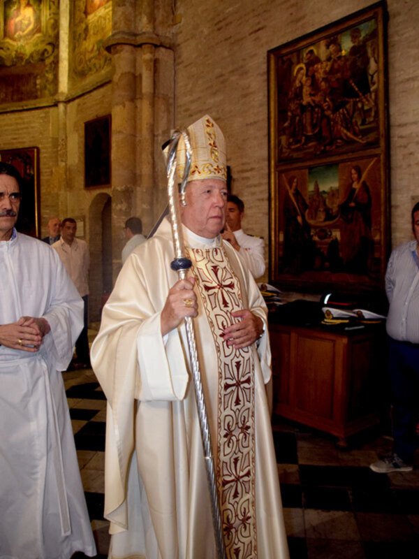 Un hombre vestido con un hábito religioso y un cetro, posiblemente un obispo, en una iglesia con pinturas en el fondo.