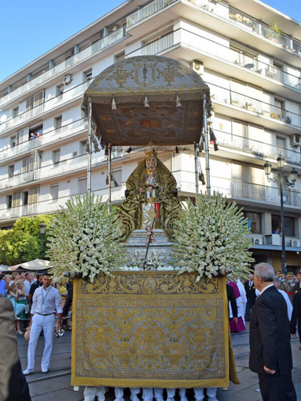 Procesión religiosa con una imagen de la Virgen María en un carro ricamente decorado, rodeada de fieles y oficiales religiosos en un entorno urbano.
