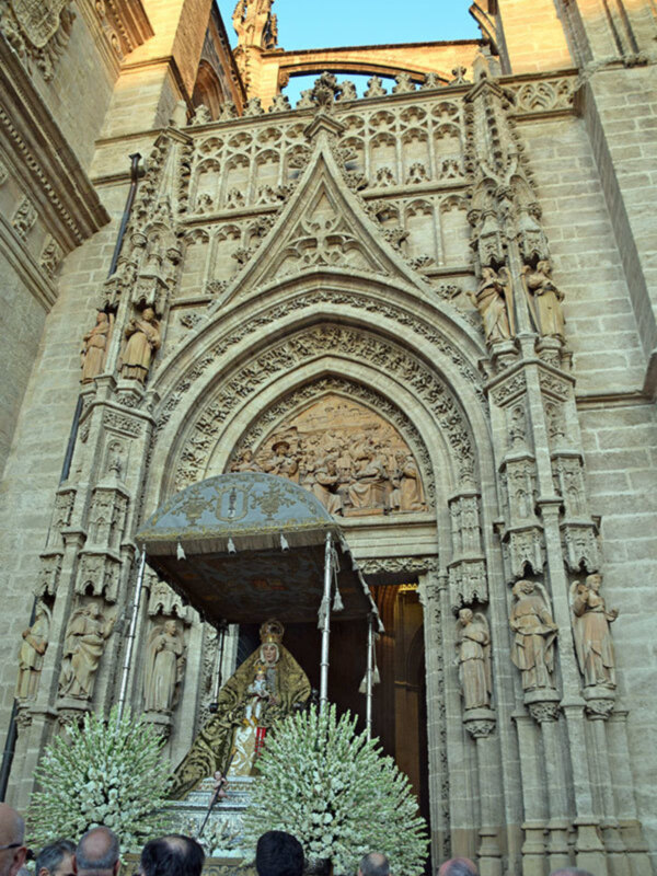 Entrada a la Catedral de Sevilla con una procesión religiosa, destacando la belleza arquitectónica y la presencia de estatuas en el portal.