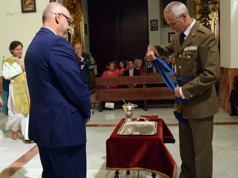 Una ceremonia en una iglesia con un militar entregando un pañuelo a un hombre vestido de traje azul.